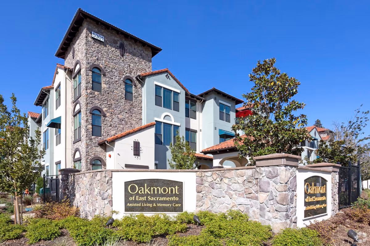 Exterior view of Oakmont of East Sacramento, a multi-story assisted living and memory care facility with stone and stucco walls, red tile roofing, and landscaped greenery in front. The building number 5301 is visible on the stone tower section.