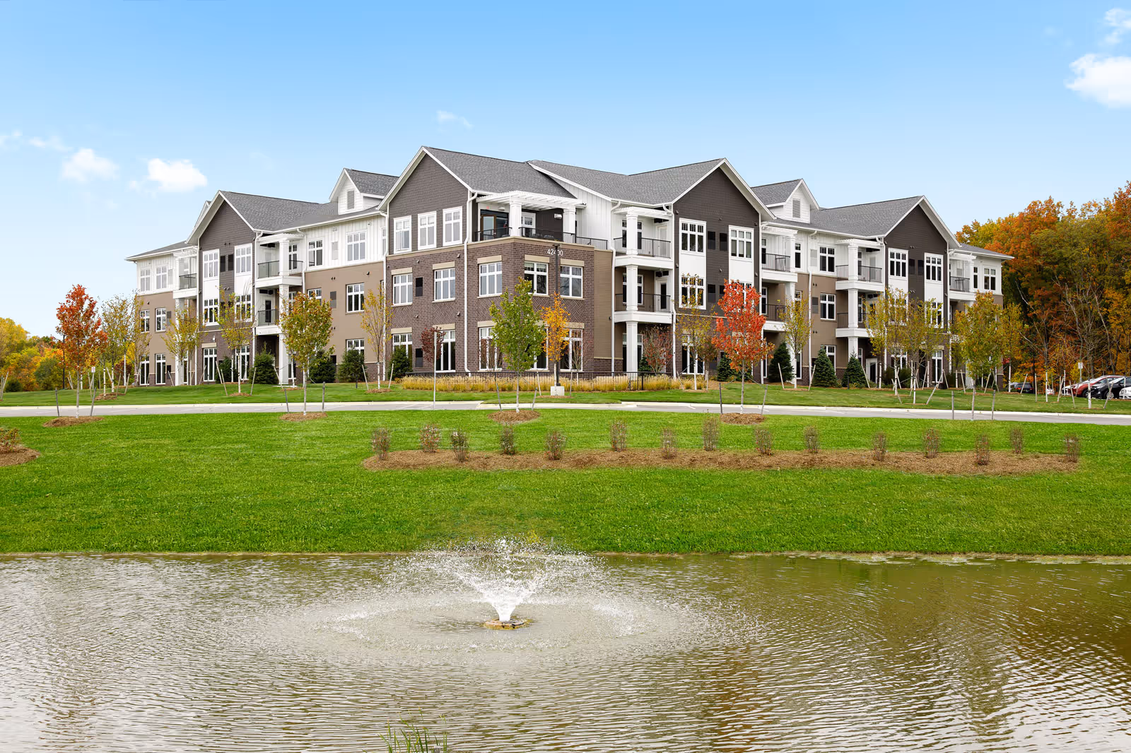 Three-story modern senior living building with balconies set behind a grassy lawn and a pond with a fountain.