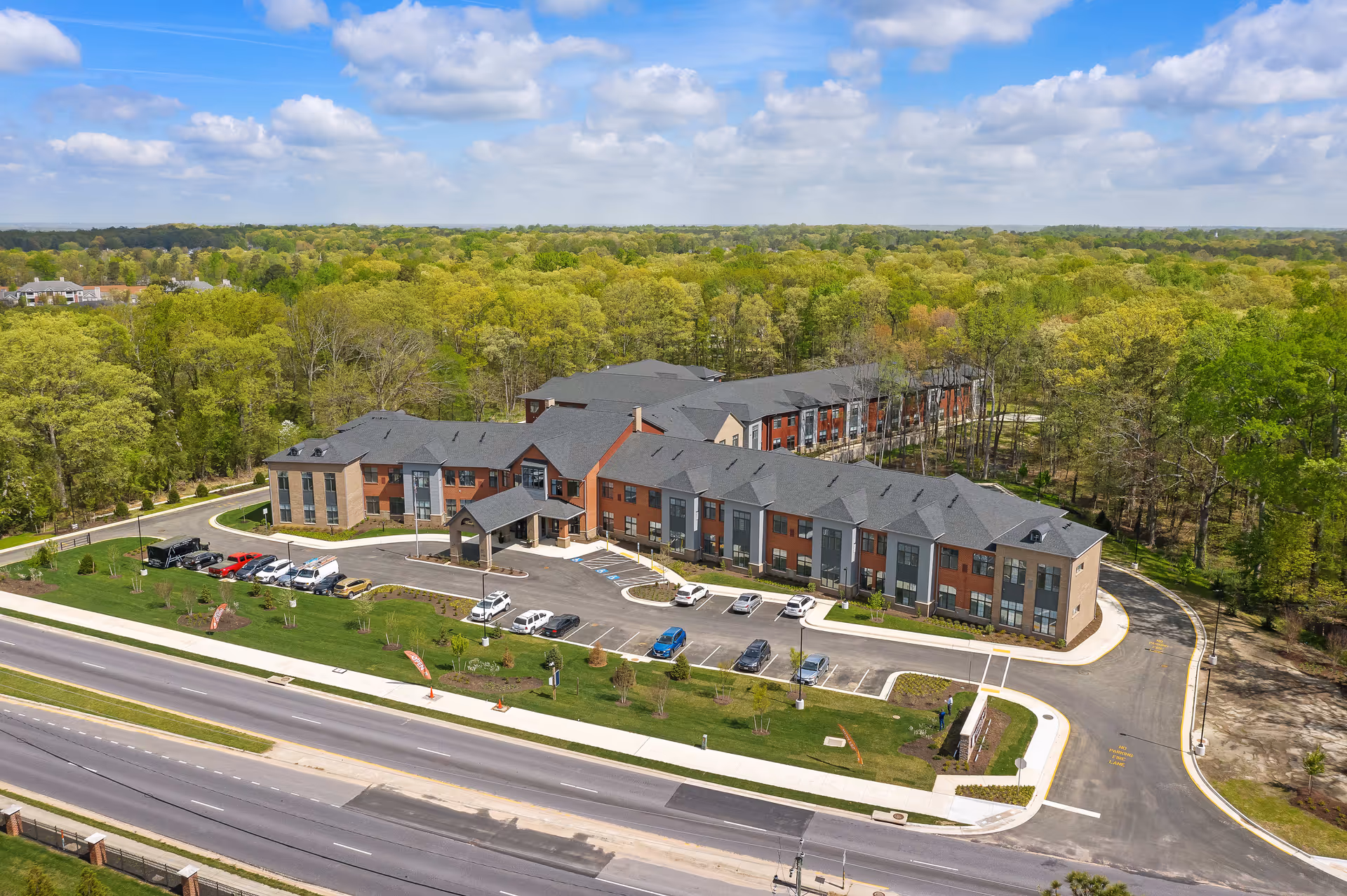 Aerial view of a three-story senior living facility with a front entrance, parking lot, and surrounding trees.