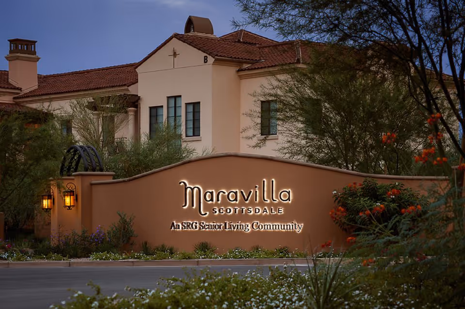 Exterior view of Maravilla Scottsdale senior living community building with a beige stucco wall and illuminated sign reading 'Maravilla Scottsdale An SRG Senior Living Community' surrounded by desert landscaping and trees at dusk.