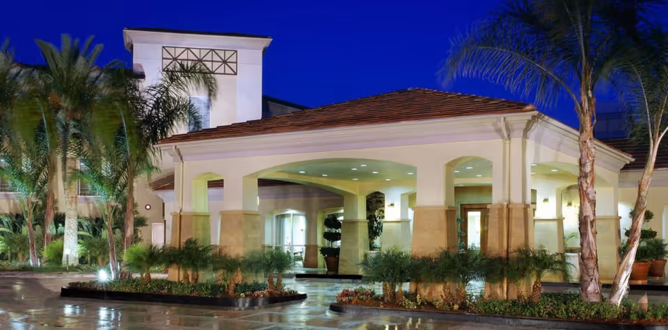Exterior view of The Village at Sherman Oaks building entrance at dusk with illuminated covered driveway, palm trees, and landscaped plants.
