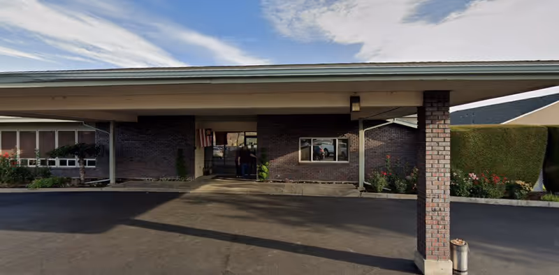Covered entrance canopy and brick front facade of a senior living facility with a driveway and landscaping beds.