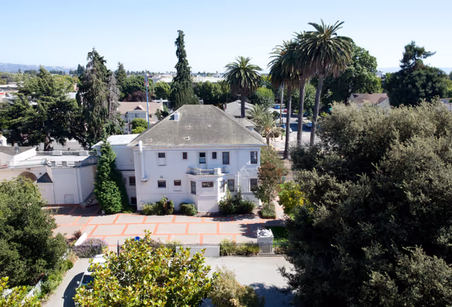 Aerial view of a white multi-story residential building surrounded by trees, palm trees, and a brick-paved courtyard.