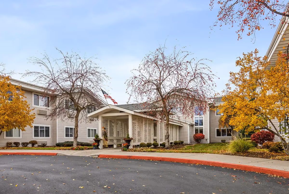 Covered front entrance of a two-story senior living building surrounded by fall foliage and a circular driveway.
