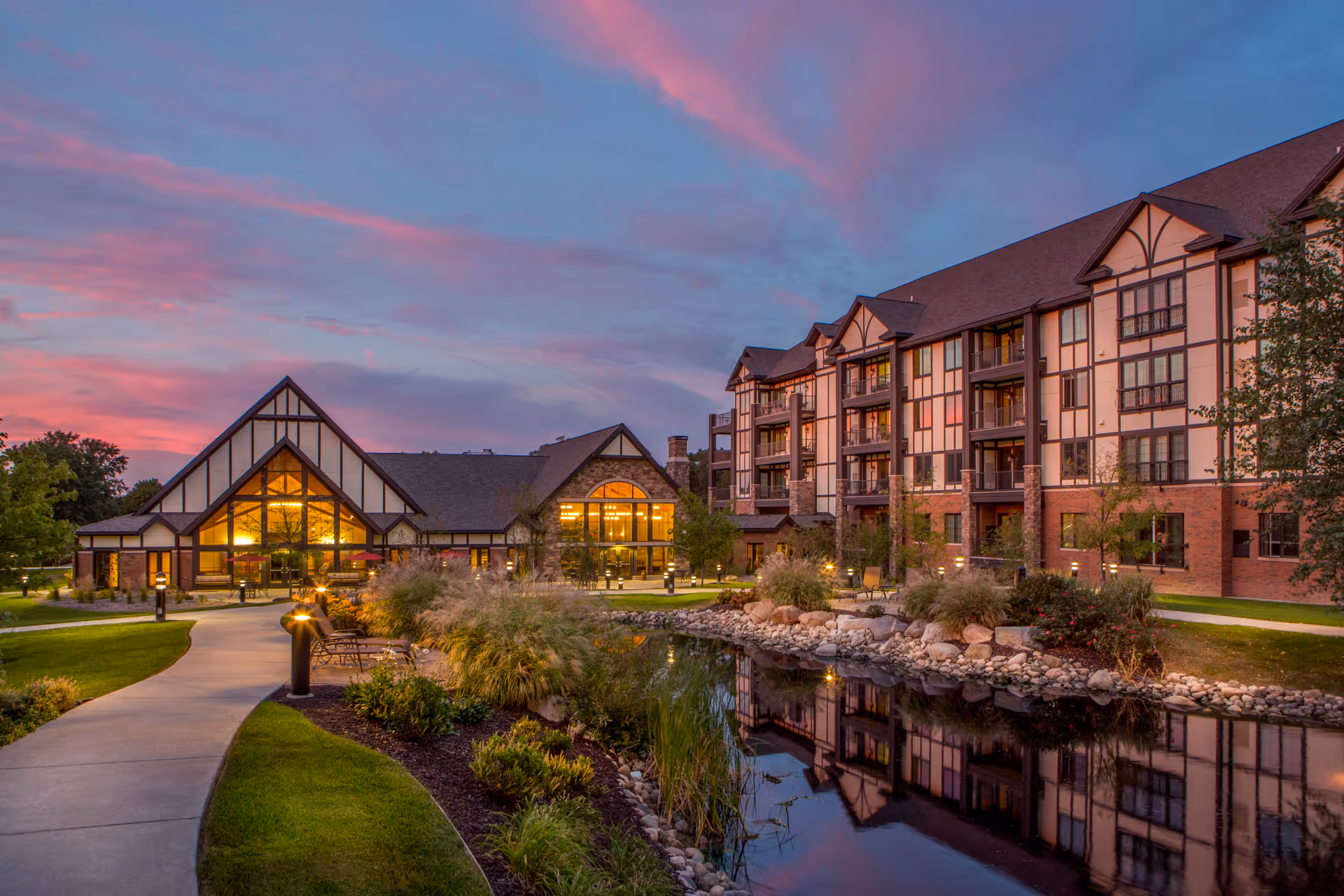 Exterior view of Beacon Hill At Eastgate facility at sunset, showing a large multi-story building with Tudor-style architecture next to a landscaped pond and walking path.