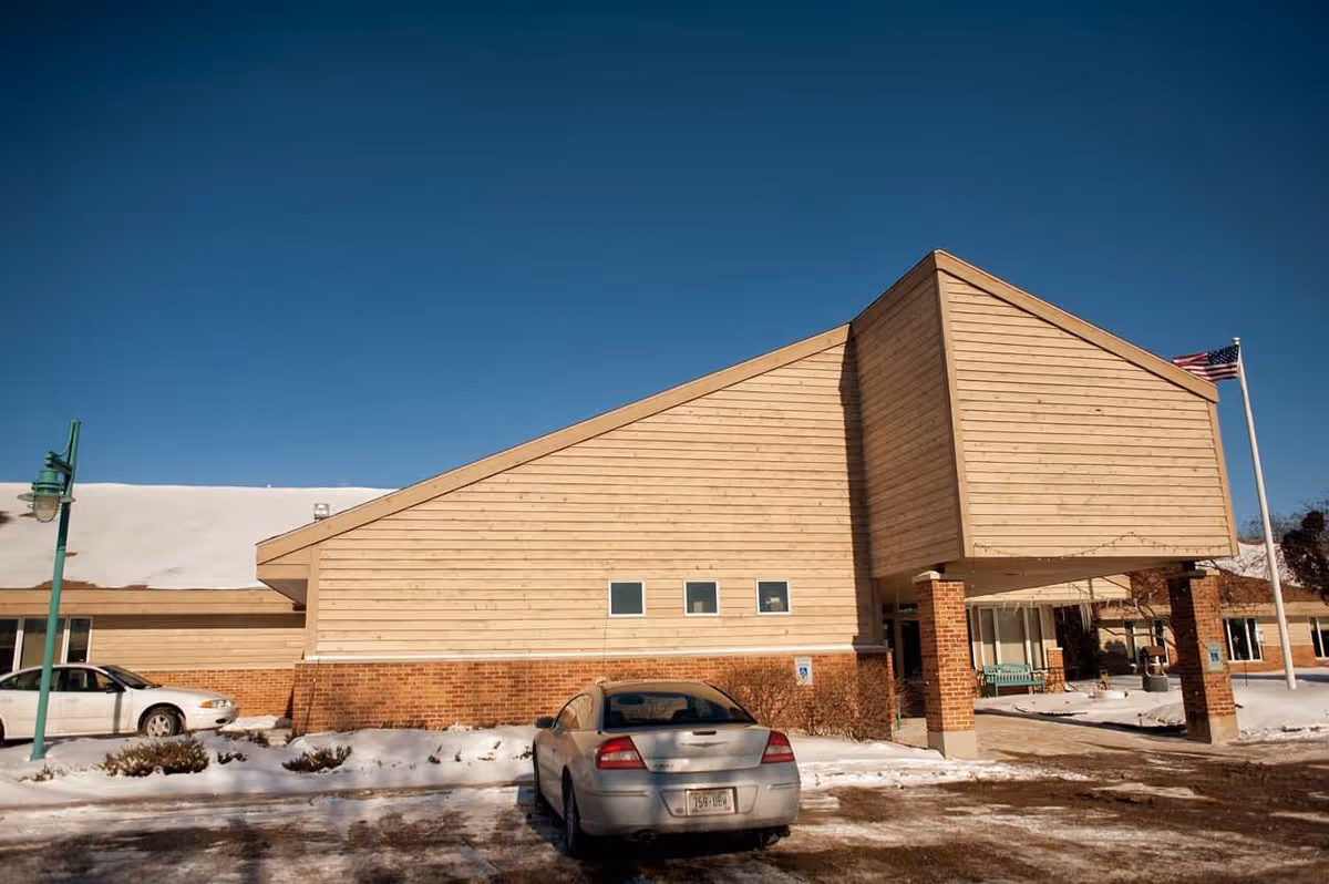 Front exterior of a single-story senior living facility with a covered entrance, parked cars, and snow on the ground.