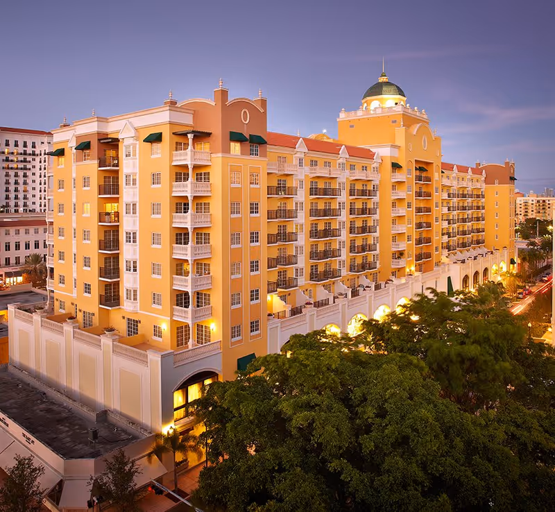 Exterior view of a large, multi-story yellow and beige building with balconies and a green dome on top, illuminated at dusk with trees in the foreground and city buildings in the background.