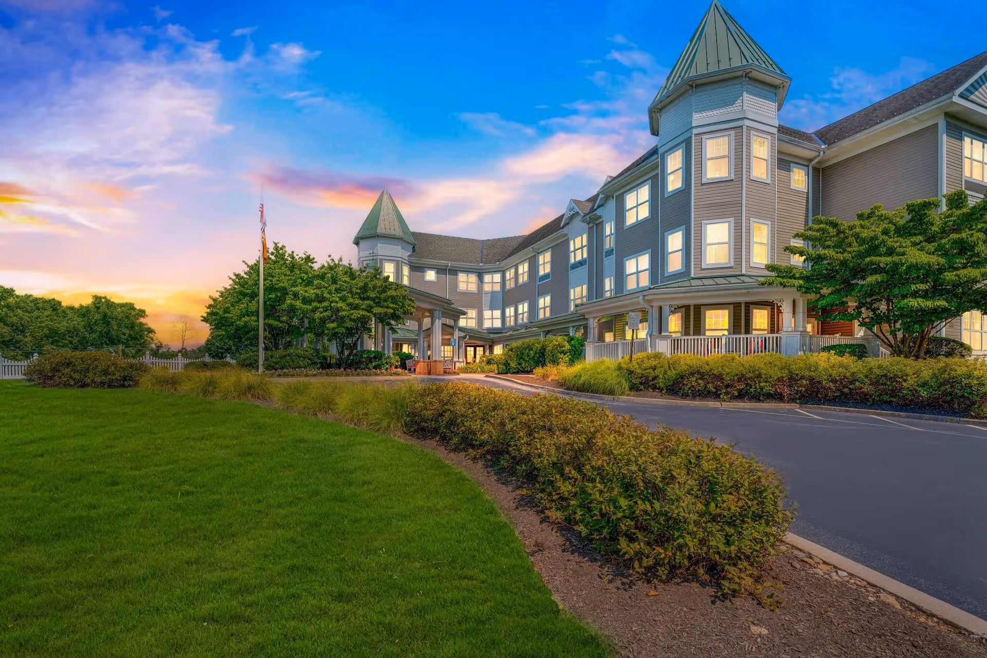 Front exterior of a multi-story senior living building at sunset with lit windows, a driveway, and landscaped lawn.