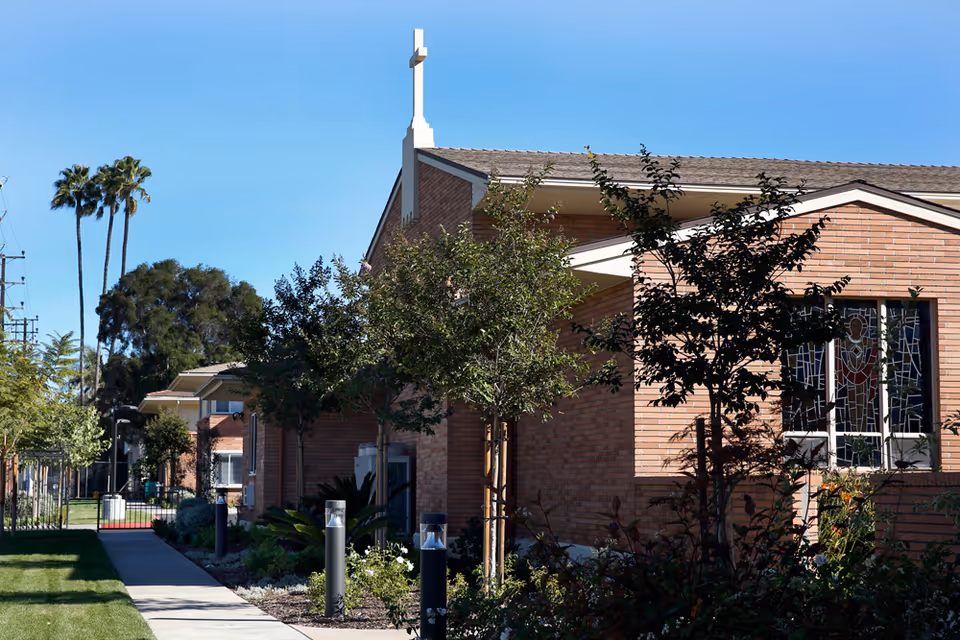 Brick church-like building with a cross on the roof, stained-glass window, trees, and a sidewalk leading past landscaped grounds.