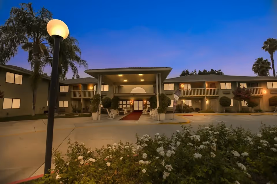 Front entrance of a two-story senior living facility at dusk with an illuminated canopy, palm trees, and flowering shrubs.