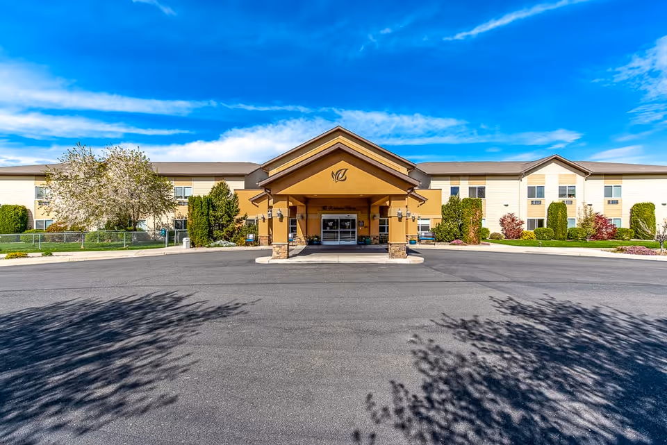 Front exterior view of Ellensburg Senior Living facility showing a two-story building with a covered entrance, surrounded by landscaped greenery and trees under a blue sky with scattered clouds.