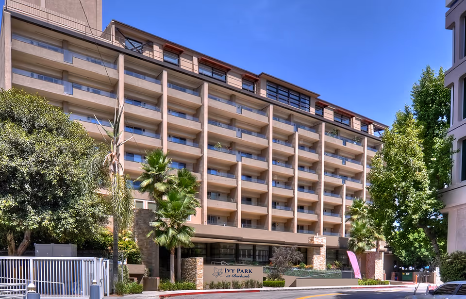 Front exterior of a multi-story senior living building with rows of balconies, palm trees, and an 'Ivy Park at Burbank' sign.