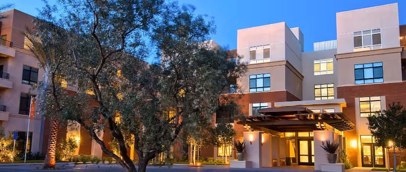 Front exterior of a modern multi-story senior living building at dusk, showing the illuminated entrance, windows, and landscaping with trees.