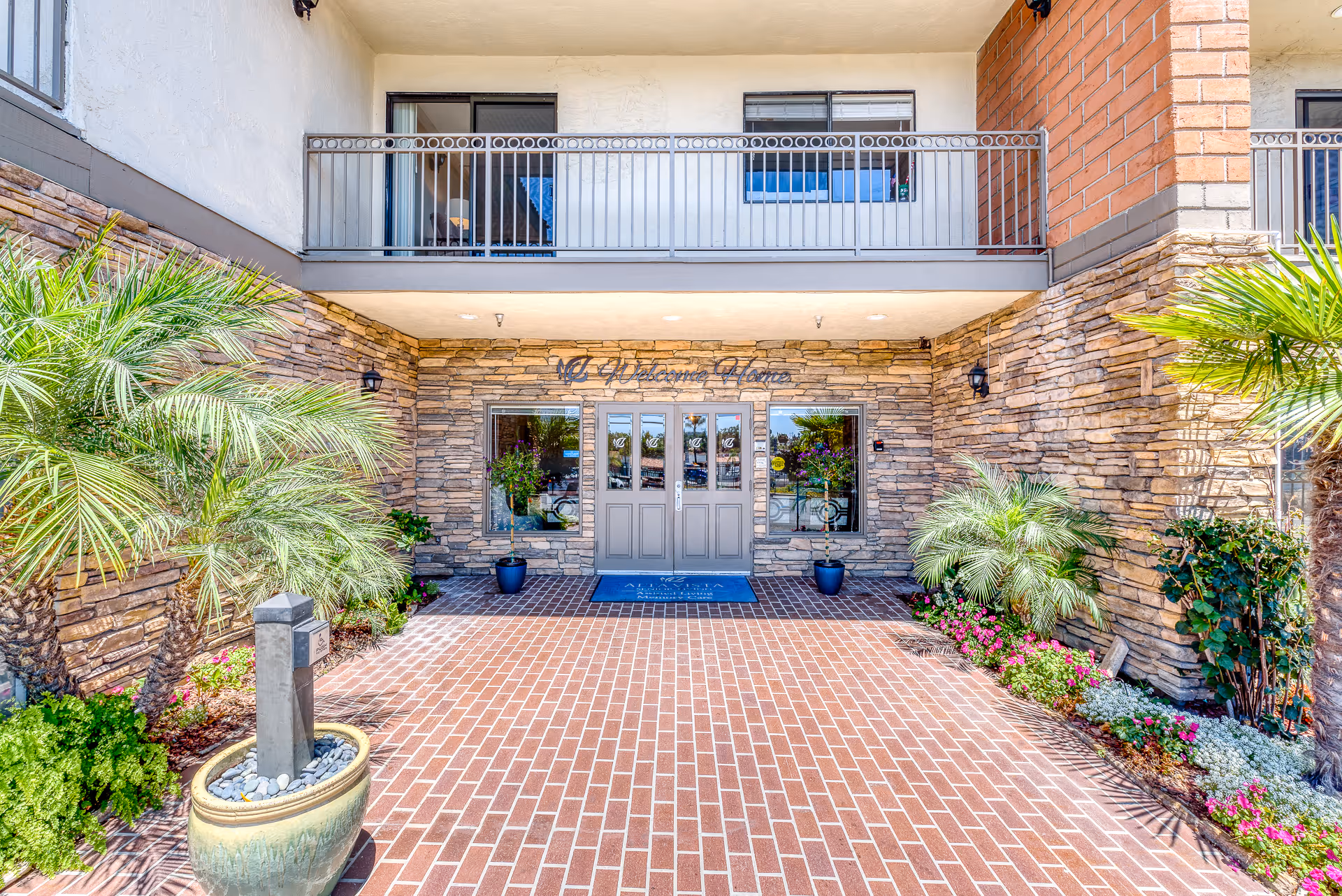 Brick-paved entrance to a senior living building with double doors set in a stone facade, a balcony above, and potted plants flanking the entry.