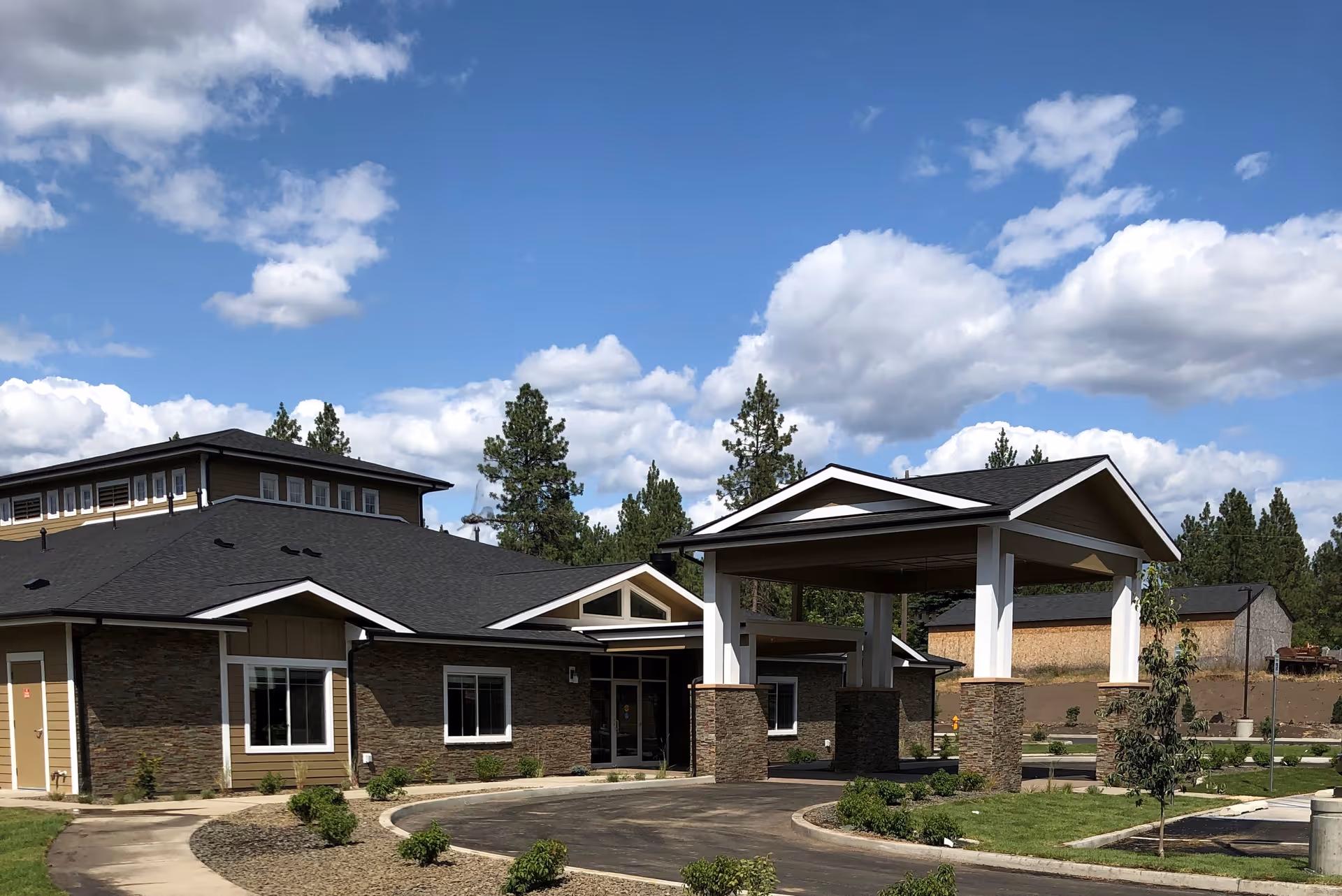 Exterior view of a single-story building with a covered entrance supported by stone pillars. The building has a dark roof, beige and stone facade, surrounded by a driveway, small landscaped bushes, and trees under a partly cloudy blue sky.