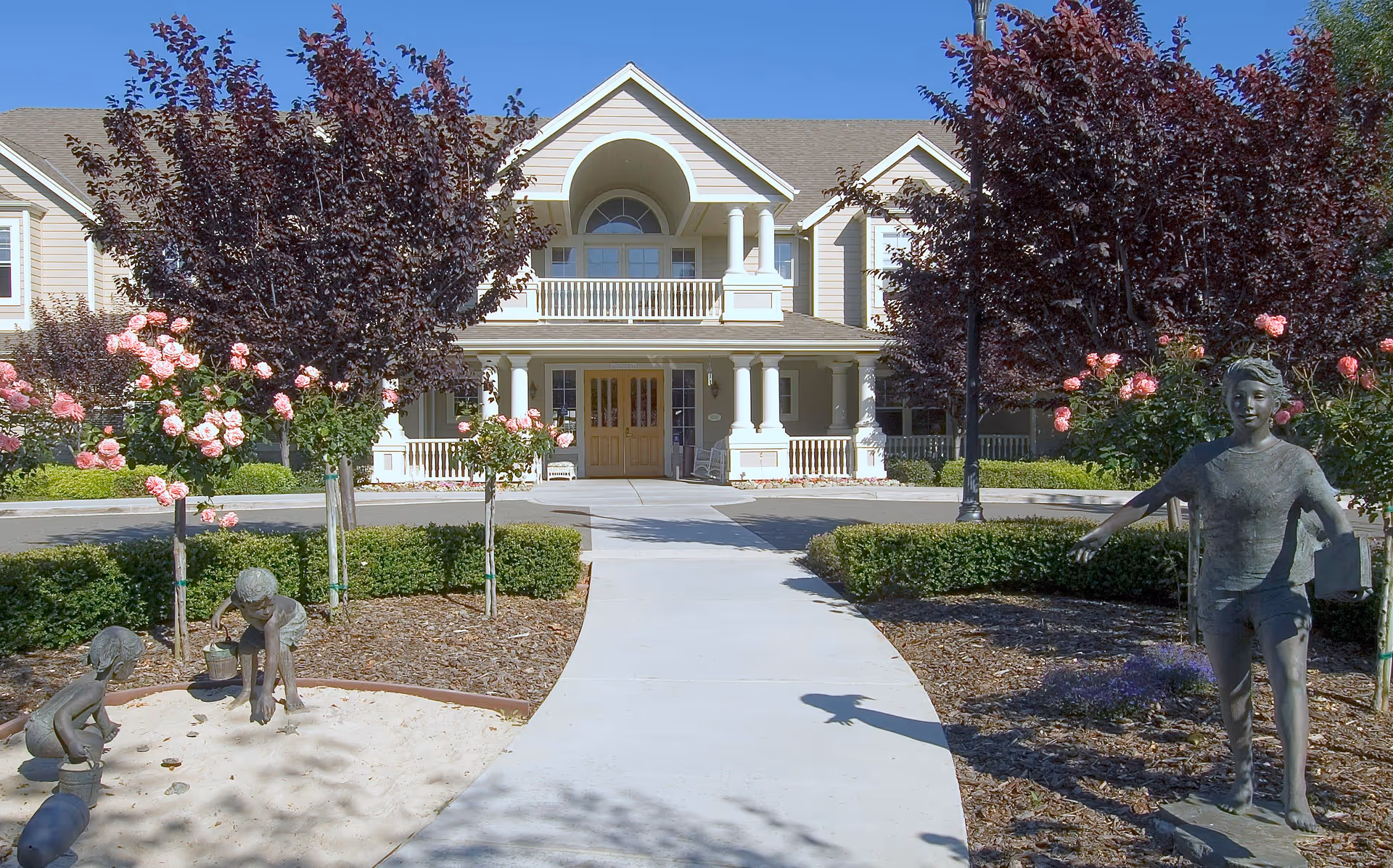 Front exterior view of a senior living facility with a wide concrete pathway leading to a covered entrance with double wooden doors. The building has beige siding, white columns, and a balcony above the entrance. The landscaped area includes pink rose bushes, small trees with dark purple leaves, and bronze statues of children playing in a sandpit and walking nearby.