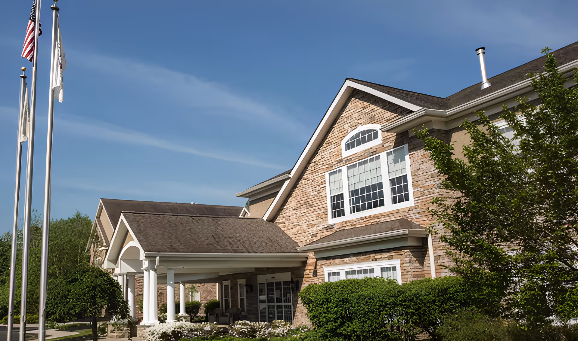 Exterior view of a senior living facility building with stone facade, large windows, and a covered entrance. There are three flagpoles with flags in front of the building, surrounded by green bushes and trees under a clear blue sky.