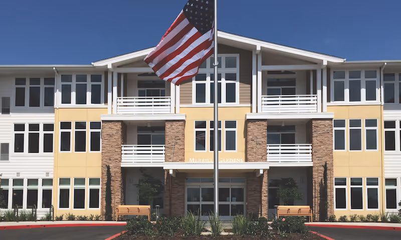 Front facade of the Merrill Gardens senior living building with balconies, an American flag on a pole in the foreground, and benches flanking the entrance.
