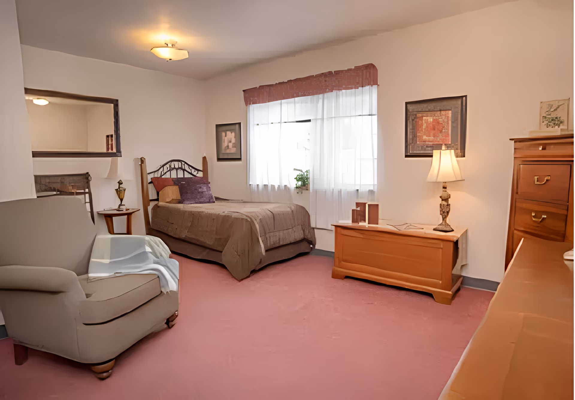 Furnished bedroom with a bed, armchair, wooden chest and dresser, lamp, and a curtained window.