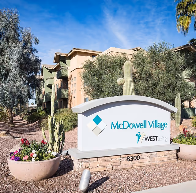 Entrance sign reading 'McDowell Village West Wing' amid desert landscaping with cacti and the apartment building under a blue sky.