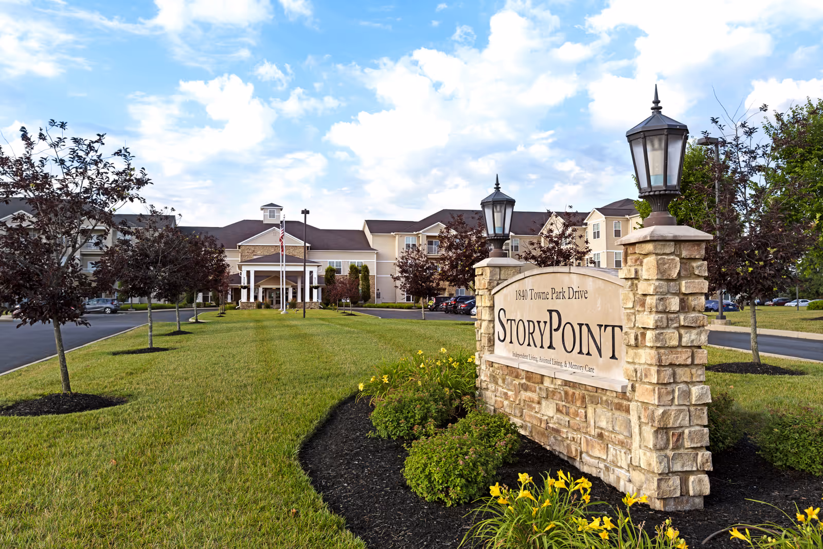 Exterior view of StoryPoint Rockford senior living facility with a stone sign in the foreground displaying the name and address. The building is in the background with a driveway, trees, and a well-maintained lawn under a partly cloudy sky.