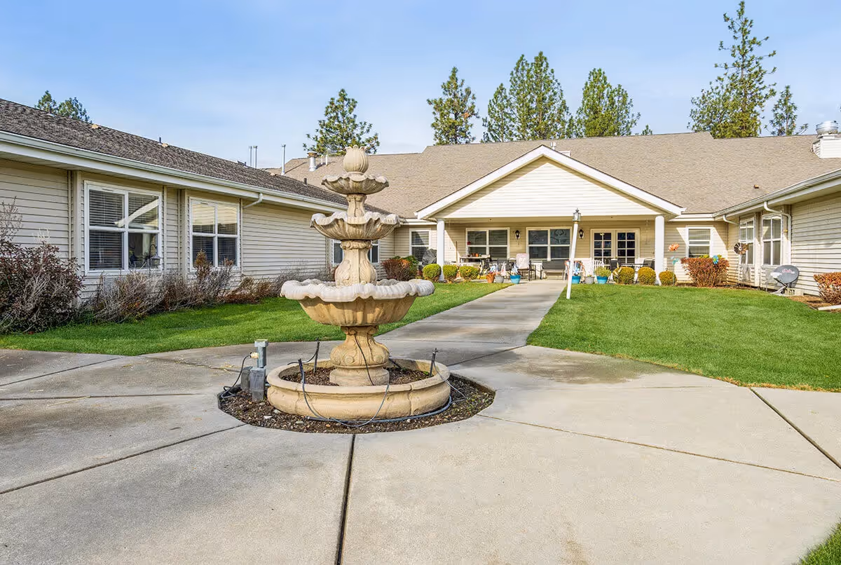 Courtyard of a single-story senior living facility featuring a central tiered stone fountain, paved walkways, green lawn, and a covered entrance porch.
