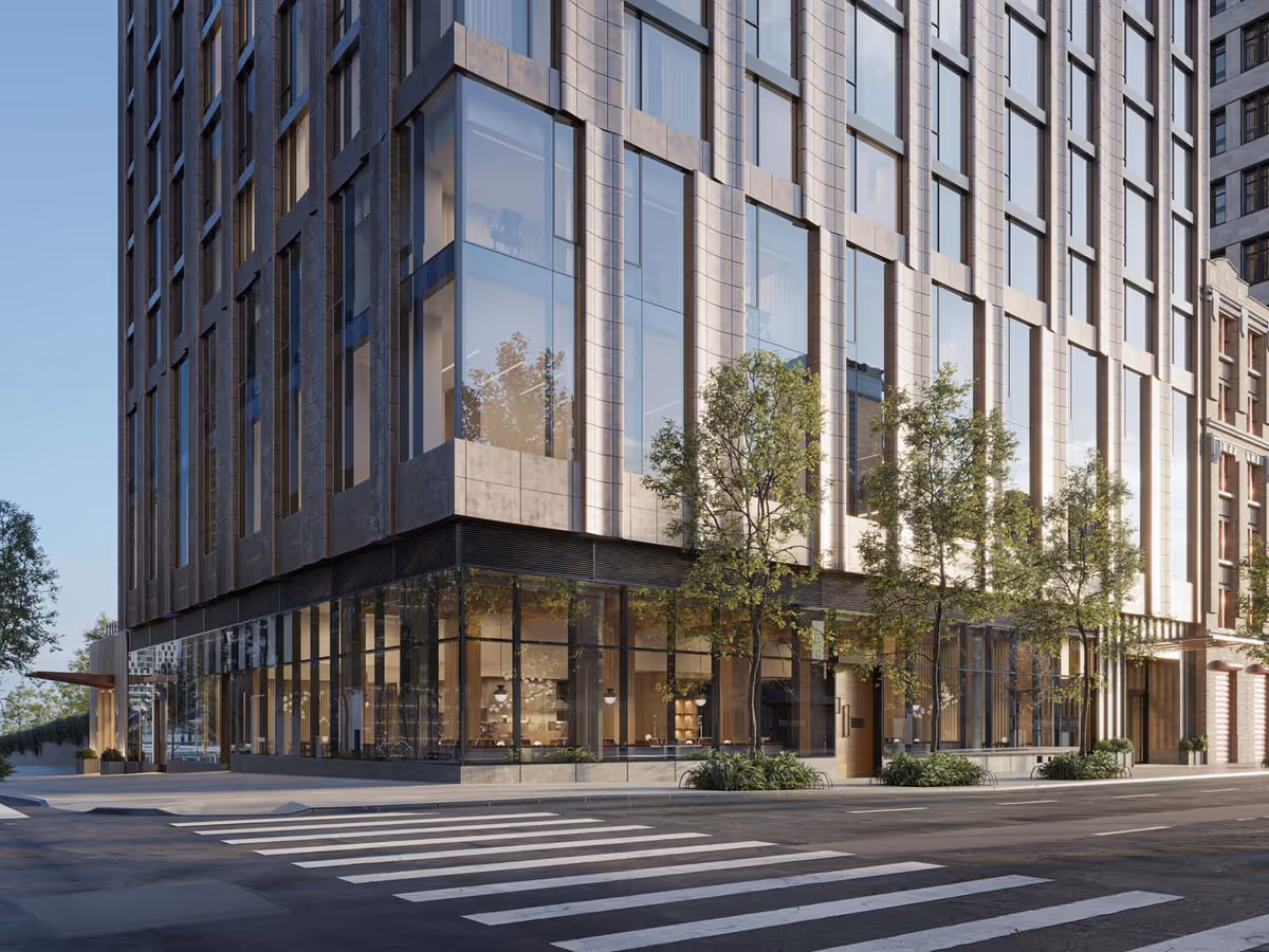 Street-level view of a modern multi-story glass-and-stone building corner with large windows, trees along the sidewalk, and a crosswalk in the foreground.