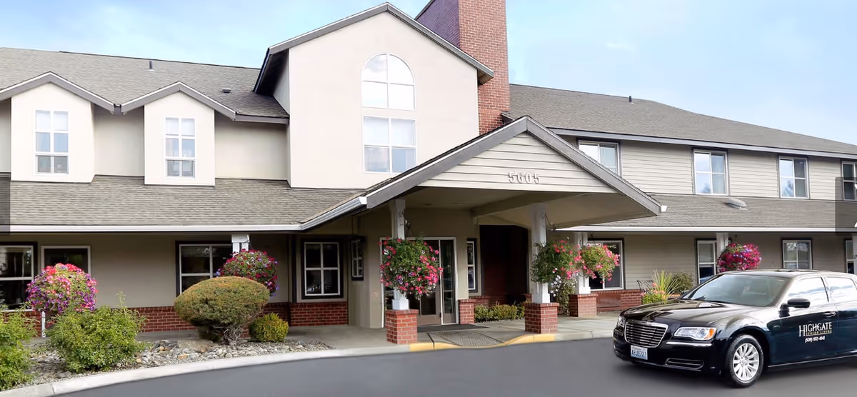 Exterior front entrance of Highgate Senior Living - Yakima with a covered porte-cochere, hanging flower baskets, landscaping, and a branded black car parked at the curb.