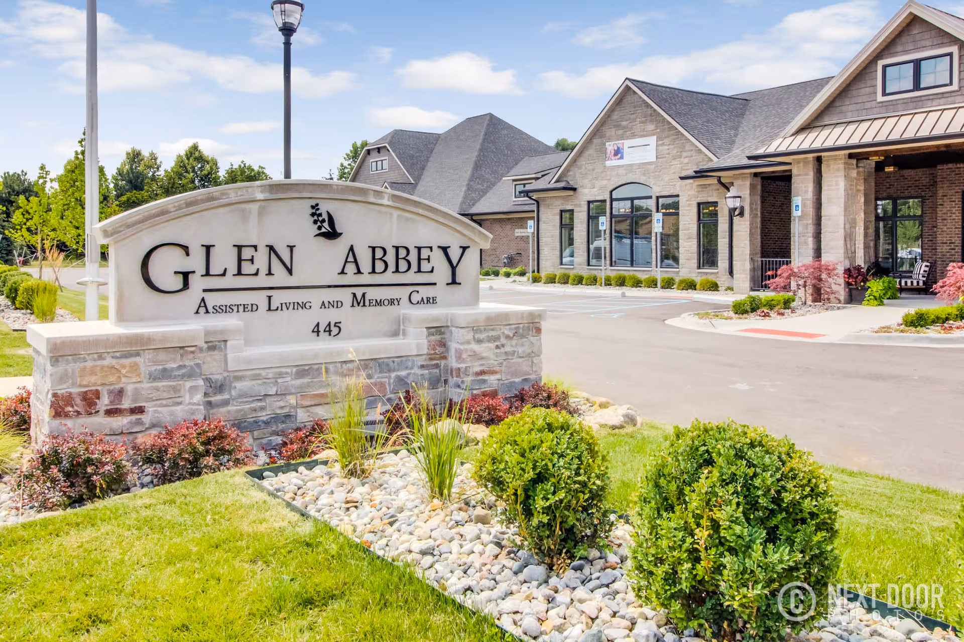 Exterior view of Glen Abbey Assisted Living and Memory Care facility with a stone sign in the foreground displaying the facility name and address number 445. The building has a brick facade with large windows and a covered entrance. Landscaping includes green grass, bushes, and a rock bed. A street lamp and a clear blue sky are visible in the background.