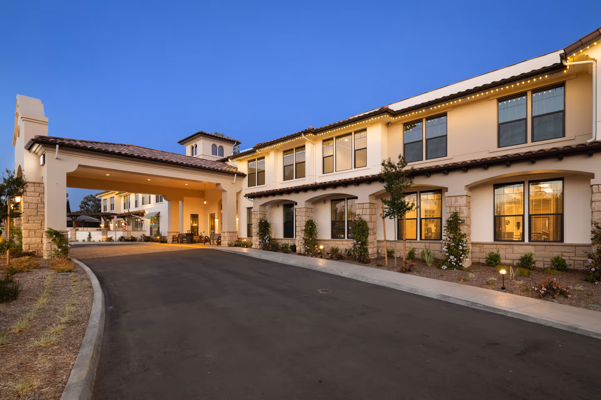 Front exterior of a two-story senior living building with a covered entryway, illuminated windows, and a paved driveway at dusk.