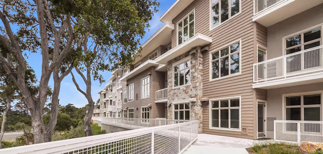 Exterior view of a modern multi-story residential building with large windows, balconies and a white metal railing walkway framed by trees.