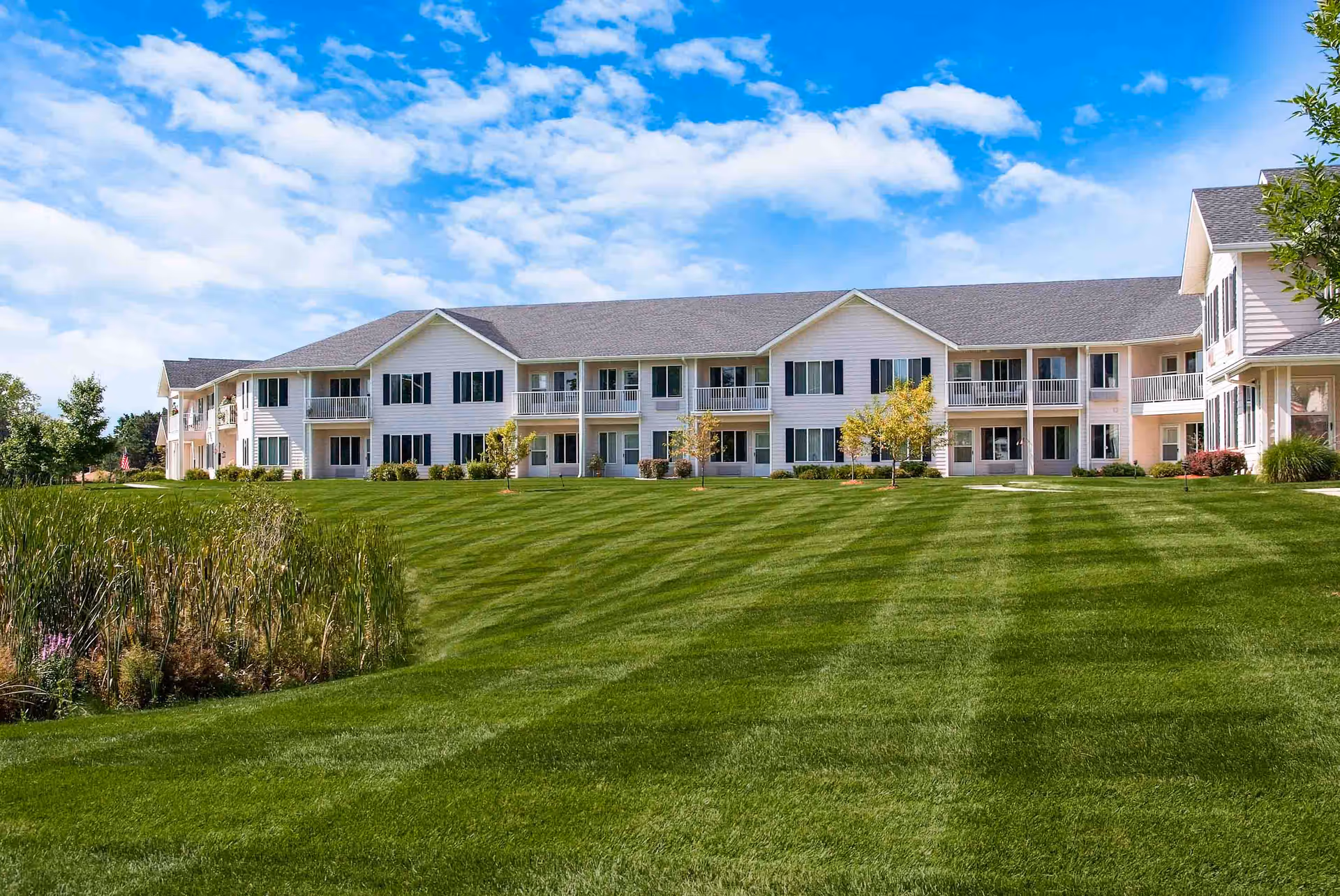 Two-story senior living building with balconies overlooking a large manicured lawn and pond under a blue sky.