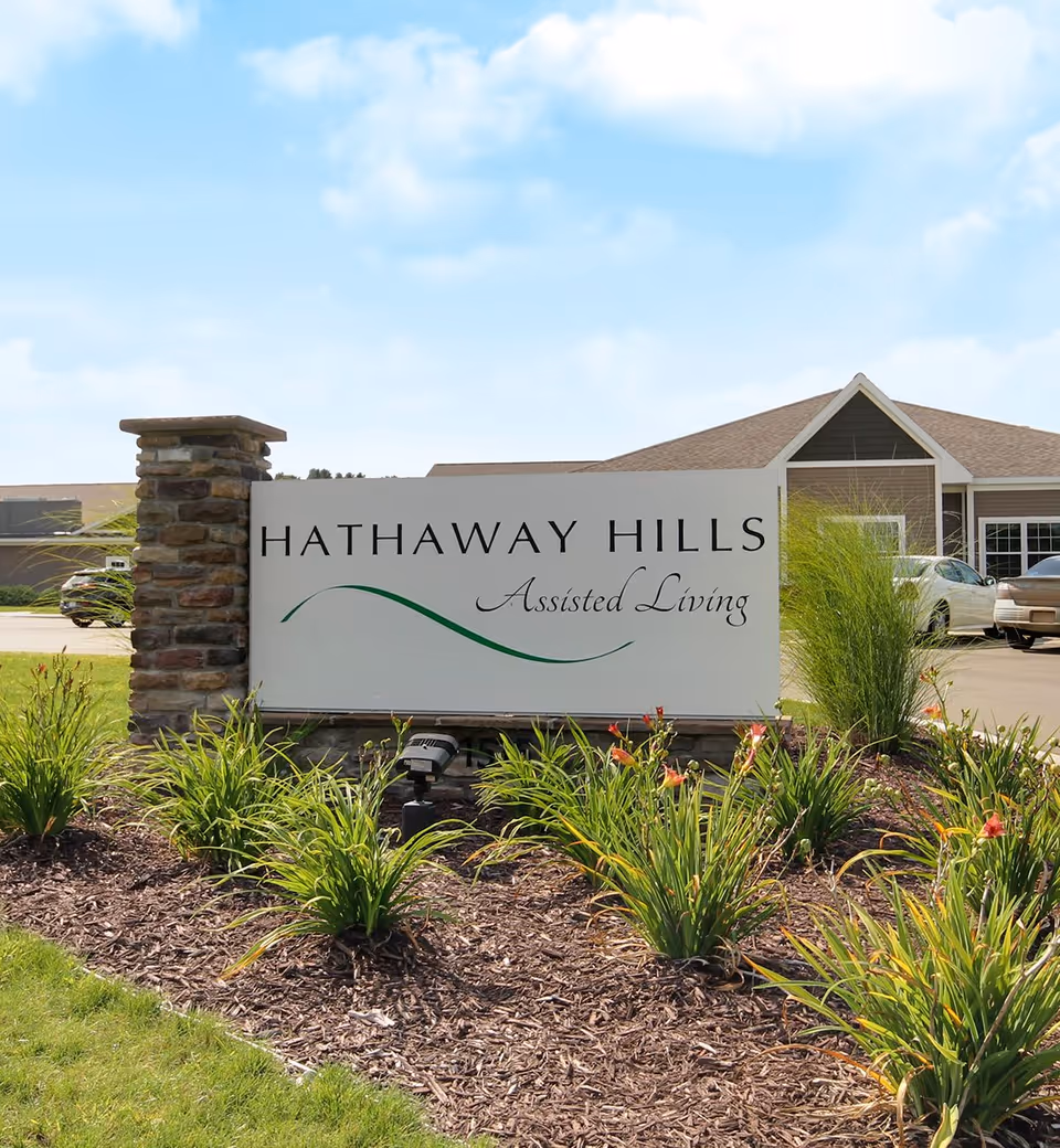 Outdoor view of a sign for Hathaway Hills Assisted Living, surrounded by landscaped plants and flowers, with a building and parked cars visible in the background under a partly cloudy sky.