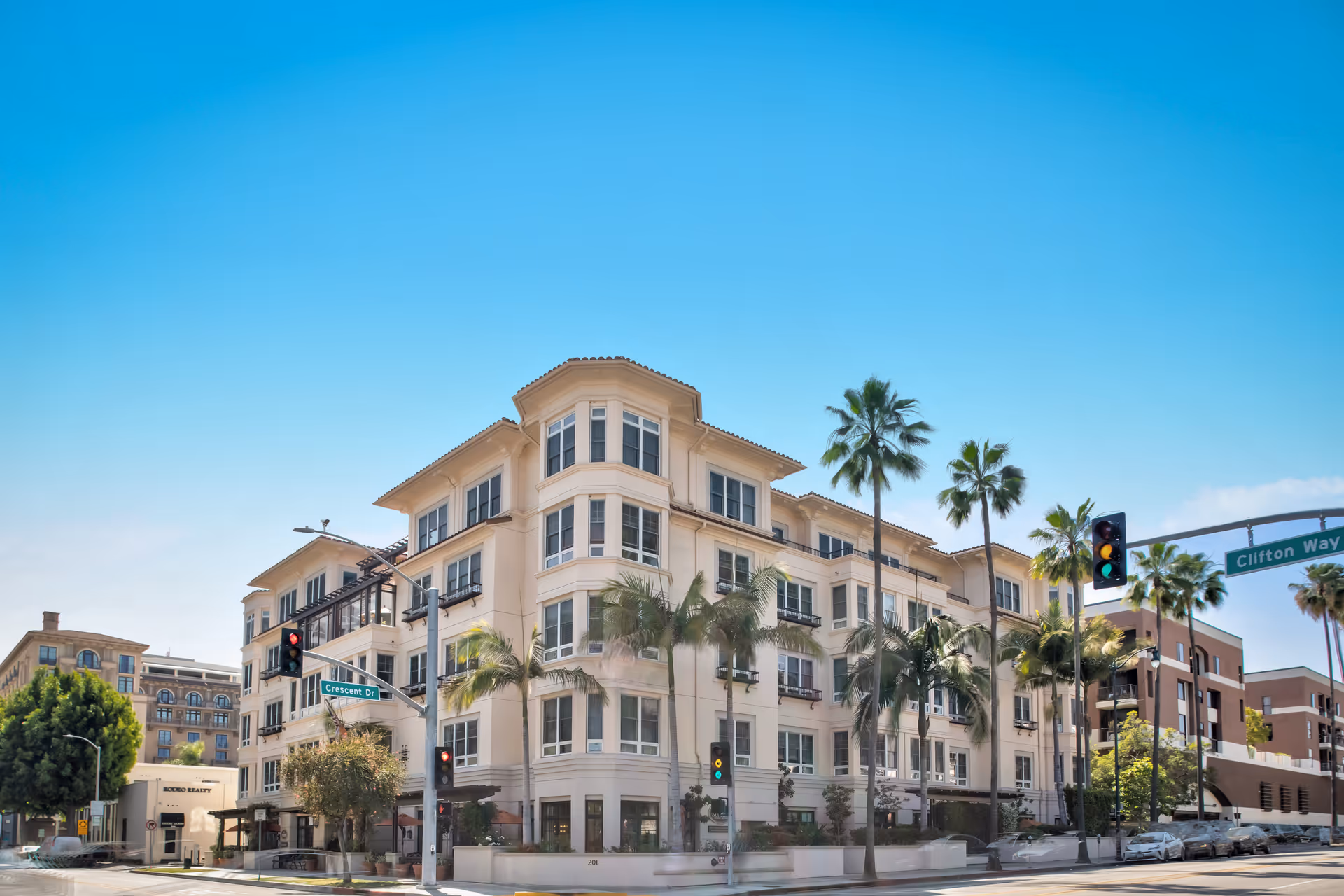 Four-story cream-colored residential building with palm trees at a street intersection under a clear blue sky.